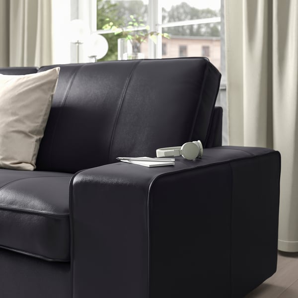 Dark KIVIK sofa with cushions, large armrests. Headphones and magazines on armrest.