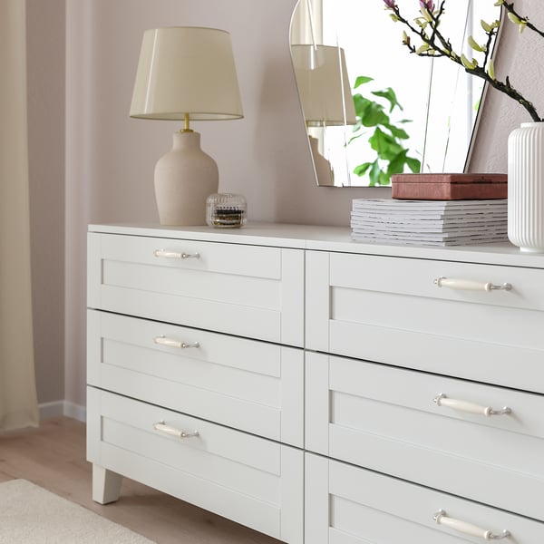White dresser with six drawers, light lamp, books, and decorative vase.