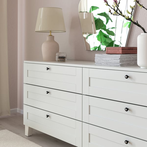 White dresser with six drawers, black knobs, lamp, vase, mirror, and books.