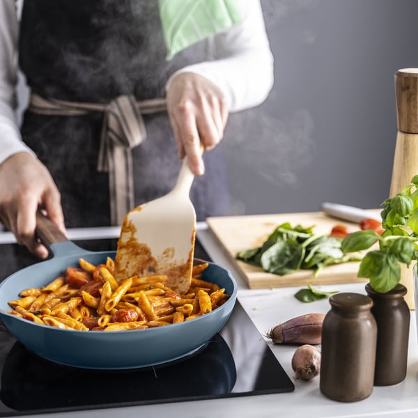 A chef stirs cooked pasta in a blue frying pan with a wooden handle, showcasing its non-stick surface and two-spout design for easy serving.