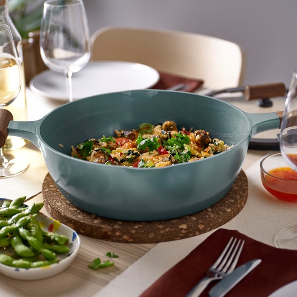 Grey-turquoise KLIPPFISK pan on table with vegetable dish. Round design, two spouts, wooden handles for serving.