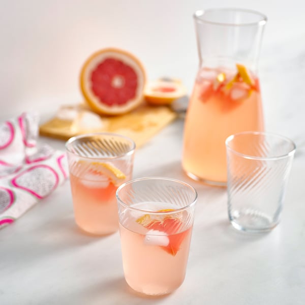 KLIPPLAX glass set: three pink drinks in thin, ribbed glasses beside a larger pitcher, halved grapefruit, and patterned napkins on a wooden board.