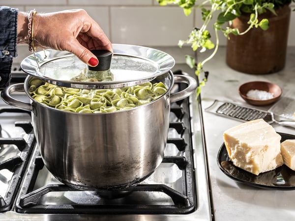 Cooking scene with KLOCKREN lid, clear glass, silver rim, on stove. Cheese and herbs nearby.