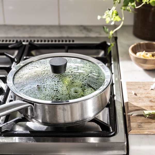 Stainless steel pan with black lid cooks greens, glass cover shows progress.
