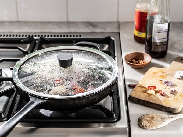 A covered frying pan on a stove. Nearby, two bottles and chopped vegetables on a wooden board.