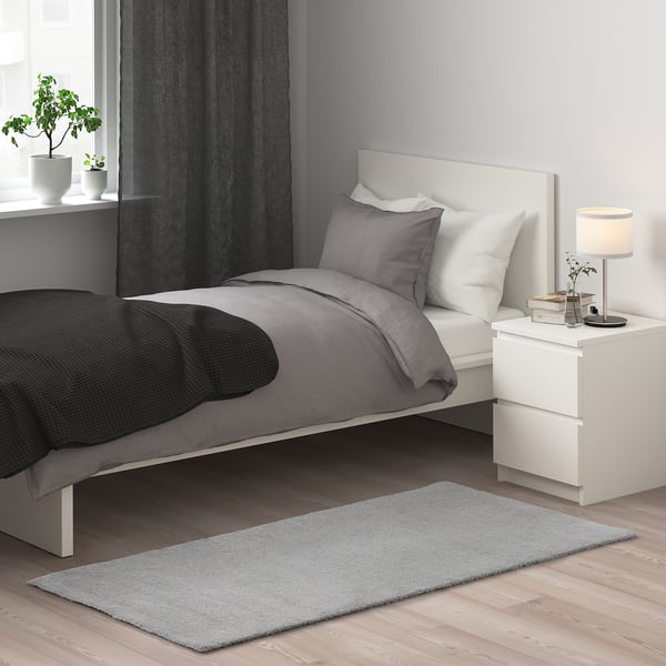 Small grey KNARDRUP rug, soft and sustainable, fits near clean white bed, contrasts dark blanket, adds warmth.