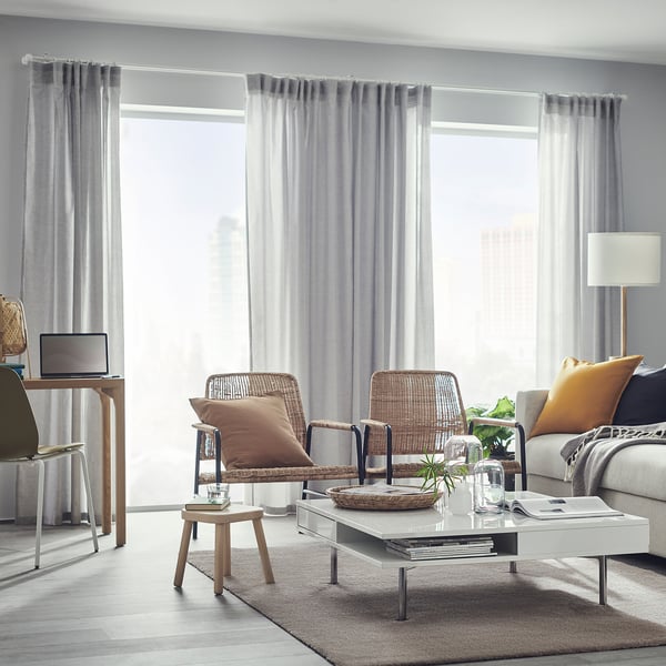 Modern living room with grey rug, wicker chairs, and glass coffee table by large windows.