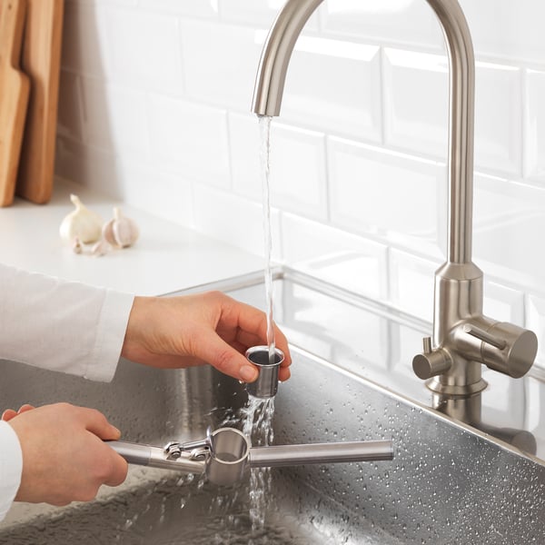 Person fills small metal cup under running kitchen tap, preparing to use it with garlic press.
