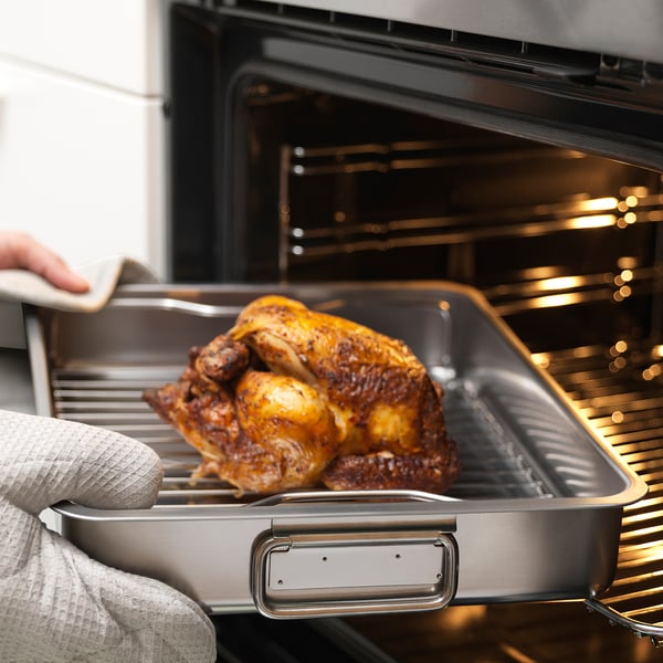 A person places a roasted chicken on a stainless steel roasting tin with a grill rack into an oven.