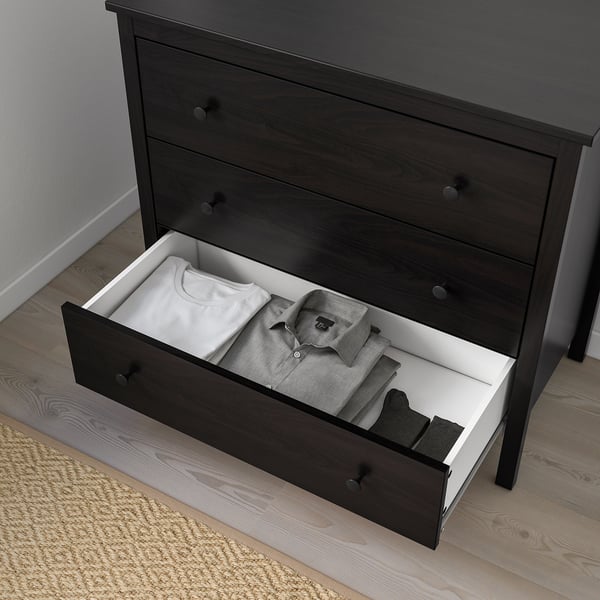 Dark wood chest of drawers with one open drawer revealing neatly folded clothes. The drawer has white lining and knobs on the front.