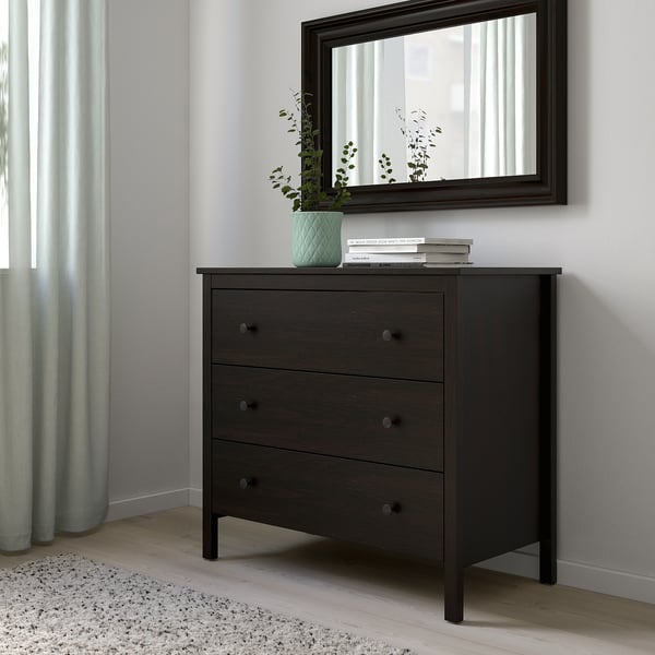 KOPPANG chest with mirror, 3 drawers, plant, books. Dark brown, fibreboard, wall-anchored. Light rug, blue curtains.