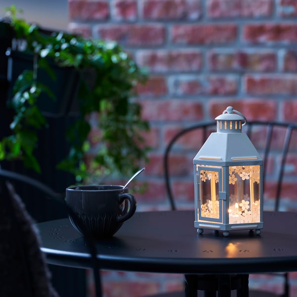 Small lantern with lit candle sits on black table. Next to it, a dark mug with spoon. The lantern glows warmly, creating a cosy ambiance.