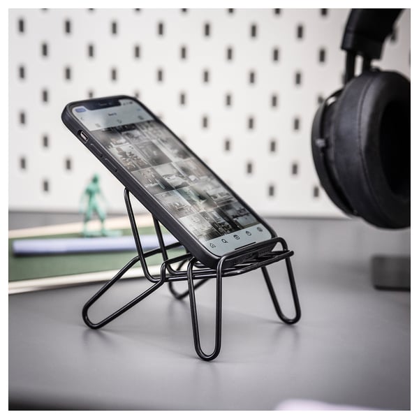 A phone stand named KRUBBET, made of black powder-coated steel, holds a smartphone. It a unique, tripod-like design.