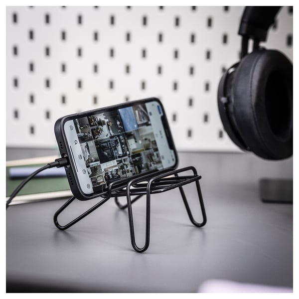 Black mobile phone stand holding a smartphone on a desk, with headphones in the background.