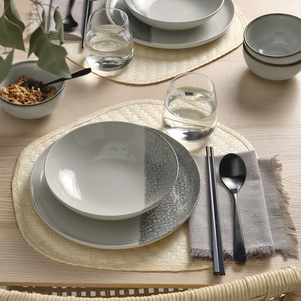 Table set with KRUSTISTEL plates, featuring grey and white designs. Includes bowls, glasses, and cutlery arranged on woven placemats.
