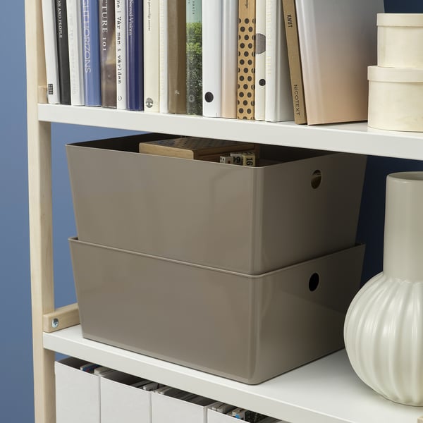 A wooden bookshelf with white shelves stores grey KUGGIS boxes and stacks books, organising items like papers or gadgets.