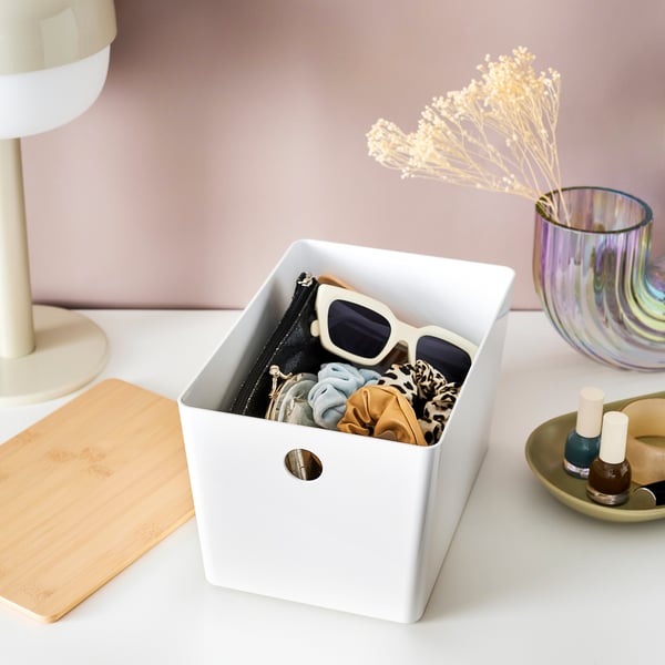 White storage box on desk holding sunglasses and scrunchies, with a lid attachment point. Also a lamp, vase, and nail polish.