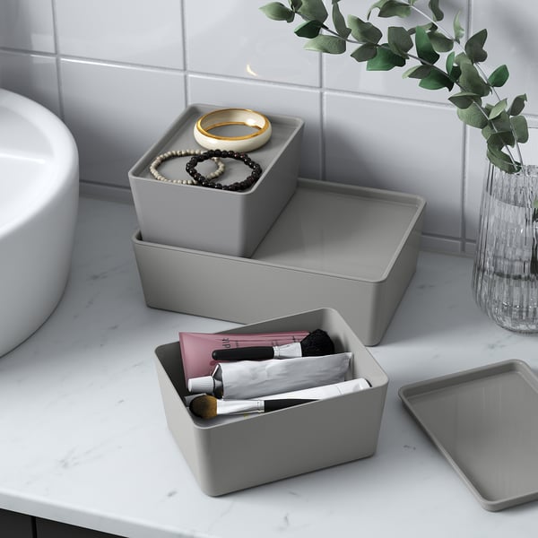 Three grey KUGGIS boxes on a bathroom counter. One holds makeup, another jewellery, and one is empty. A vase with greenery is also present.