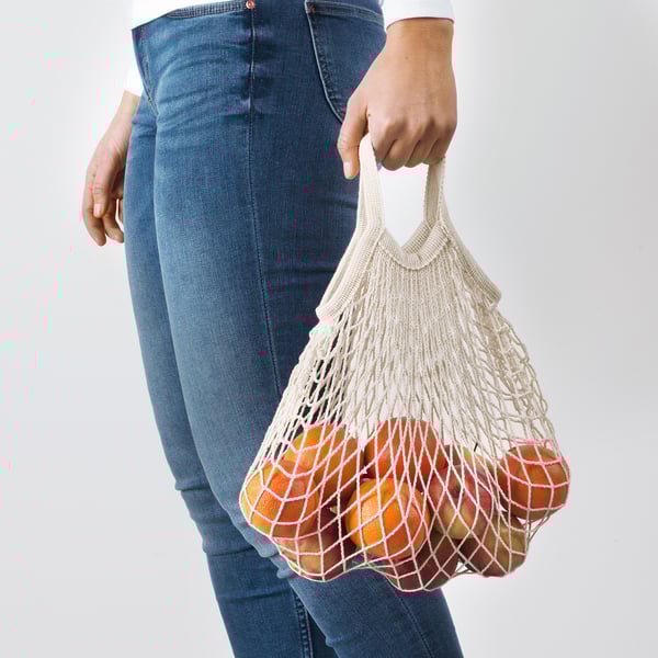 Person holding reusable net bag filled with oranges, promoting eco-friendly shopping.
