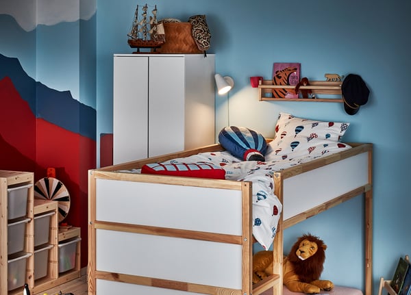 Childrens room with blue and red walls, featuring a white KURA wooden bunk bed with colourful bedding and stuffed lion.