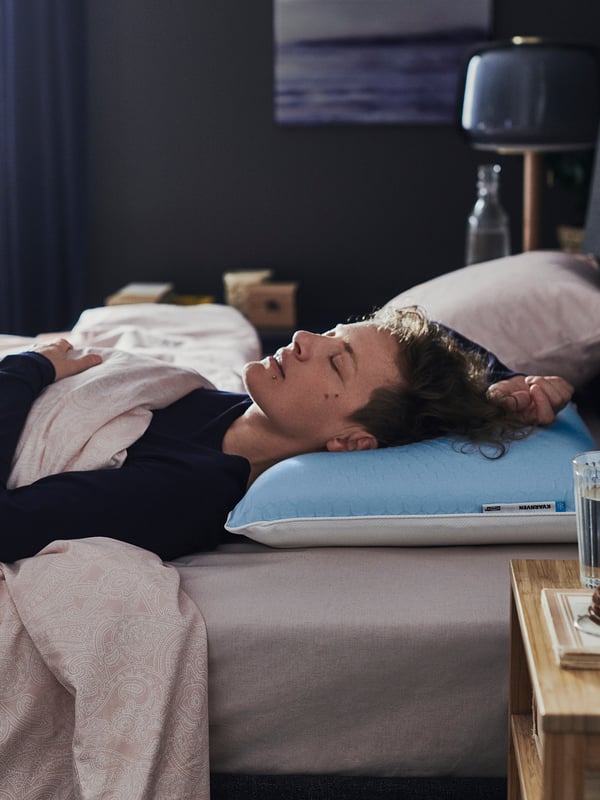 A person sleeping on a blue and white ergonomic pillow by KVARNVEN, with a bedside table nearby.