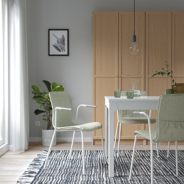 Modern dining area with grey walls, light wood cabinets, white table, 3 green chairs, black/white rug, and hanging bulb light.