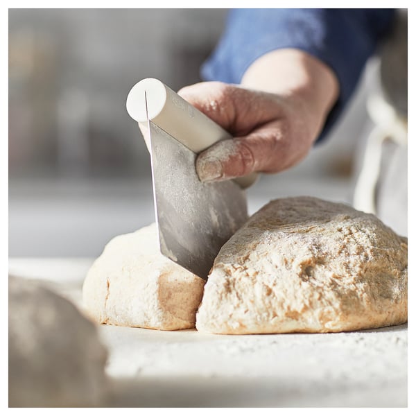 A person uses a white plastic dough cutter with a stainless steel blade to divide baked loaves on a worktop.