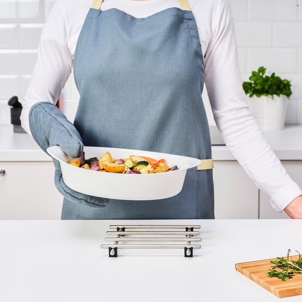 Person wearing an apron holds a white oven dish with various roasted vegetables over a counter with a metal trivet and herbs.
