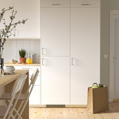 A compact white integrated kitchen freezer, slightly smaller than tall cabinets, placed beside chairs and a groceries bag.