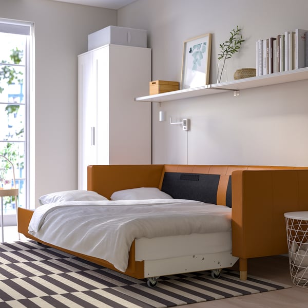 Bed frame with wheels, white bedding, wooden legs. Tall white wardrobe, window with balcony view. Striped rug, shelves with books and plant.