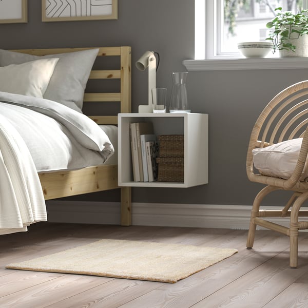 Cozy bedroom with off-white rug, wooden bed, nightstand, lamp, books, and wicker chair.