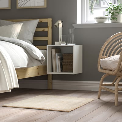 Cozy bedroom with off-white rug, wooden bed, nightstand, lamp, books, and wicker chair.