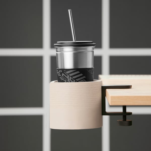 A LÅNESPELARE mug holder in light wood, clamping a silver mug with a black sleeve. The holder is mounted on a desk, keeping the drink within reach.