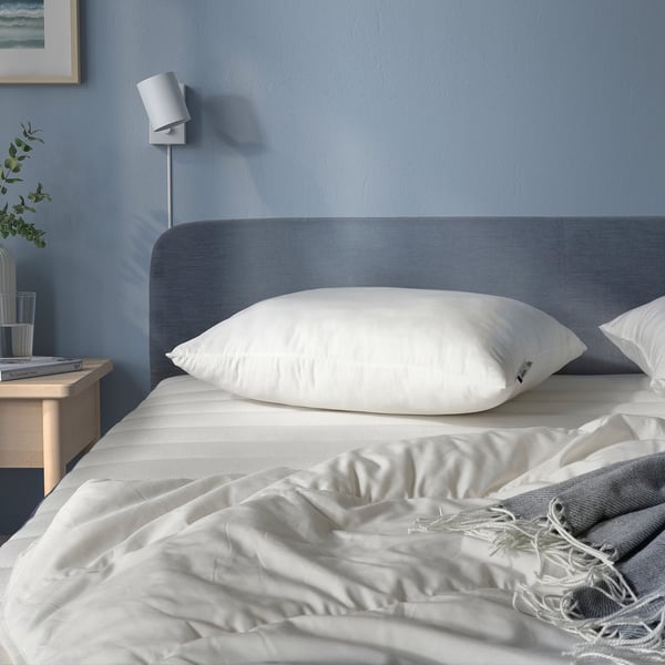 Blue bed with large white pillow, grey blanket, bedside table, plant, lamp.