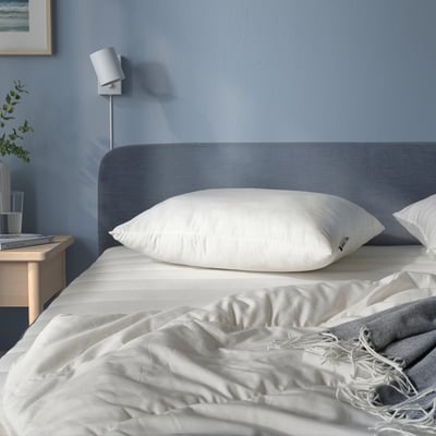 Blue bed with large white pillow, grey blanket, bedside table, plant, lamp.