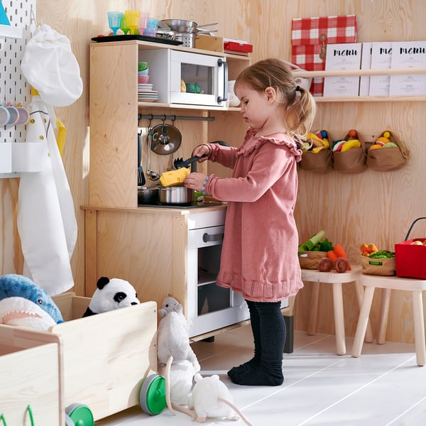 Young girl plays in wooden kitchen, with stuffed animals, colourful cups, and hanging apron, preparing food with a toy set.