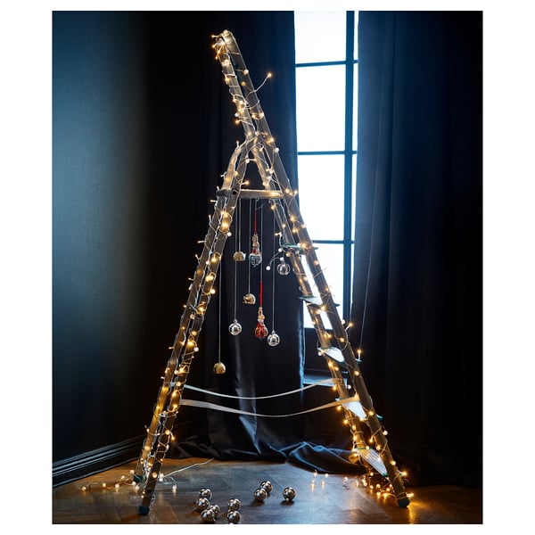 Unique triangular christmas tree with led lights and ornaments against dark curtains by window.