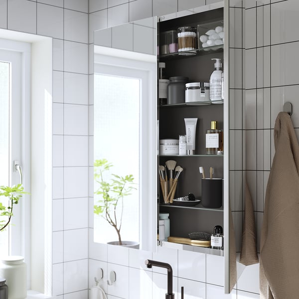 Modern wall-mounted mirror cabinet with open door, showing toiletries and cosmetics.