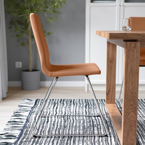 A modern chair with a brown, cushioned seat and back, supported by a chrome sled base, positioned near a wooden table with a patterned rug underneath.