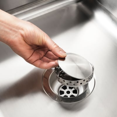 A hand adjusting a small metal sink stopper, part of the LILLVIKEN system, designed to control water flow in a kitchen sink.