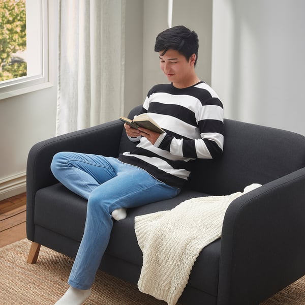 A person reads on a LINANÄS dark grey sofa, striped shirt, blue jeans, cozy blanket.