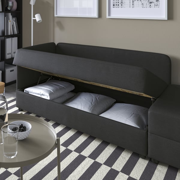Black sofa bed, partially opened to reveal storage area with pillows. Next to a round table with glasses and snacks.