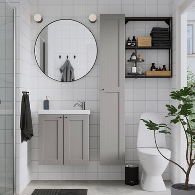 Modern bathroom: round mirror, black frame, grey vanity. White toilet, storage, plant. Glass shower.