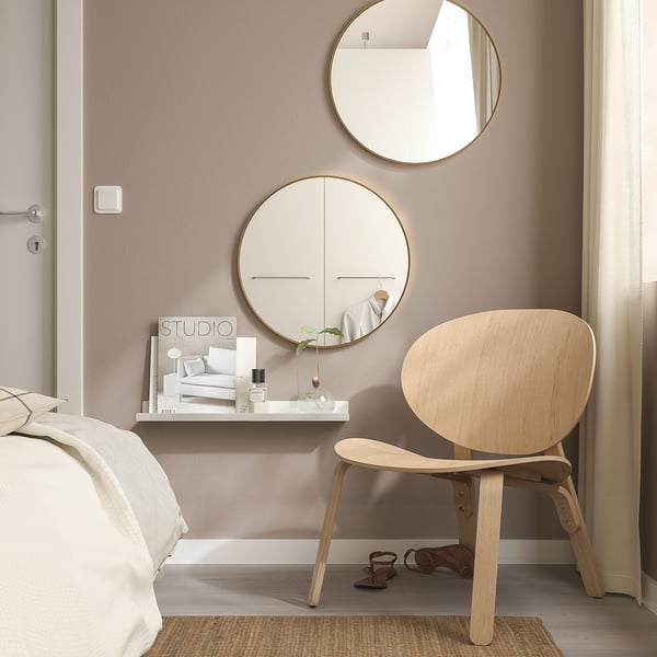Minimalist bedroom with gold LINDBYN mirror, reflecting room. Light wood chair, desk with candle, magazine, calm tones.
