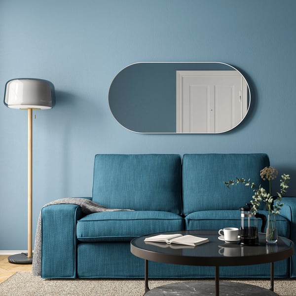 A LINDBYN white oval mirror above blue sofa, reflecting door and lamp.