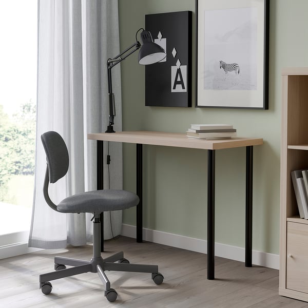 Wooden LINNMON desk with black legs, grey swivel chair, two artworks, bookshelf, and large window with sheer curtains.