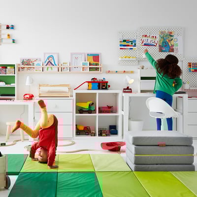 Kids playroom: two children playing with toys. One does handstand on green mat, other on cushions. Modern desk and adjustable chair.