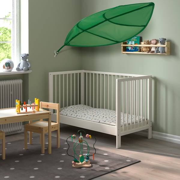Green leaf canopy hangs above white crib in cosy nursery with sage walls, wooden furniture, and toys.