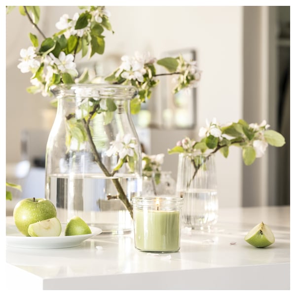 In a green apple candle in a glass sits on a kitchen counter, surrounded by cut apples, white flowers in vases, and a plate. Elegant and fresh decor.