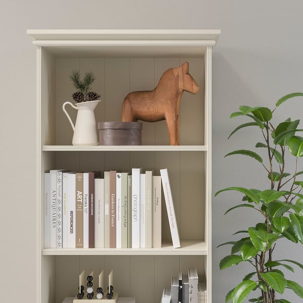 A white bookcase with adjustable shelves, displaying books, a wooden horse, and a plant.